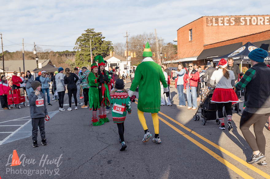 Elf-costumed volunteers greet a Buddy the Elf runner and a child on a sunlit street corner