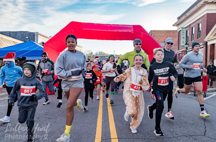 Children and adults in festive costumes run toward the camera out from under the red start arch